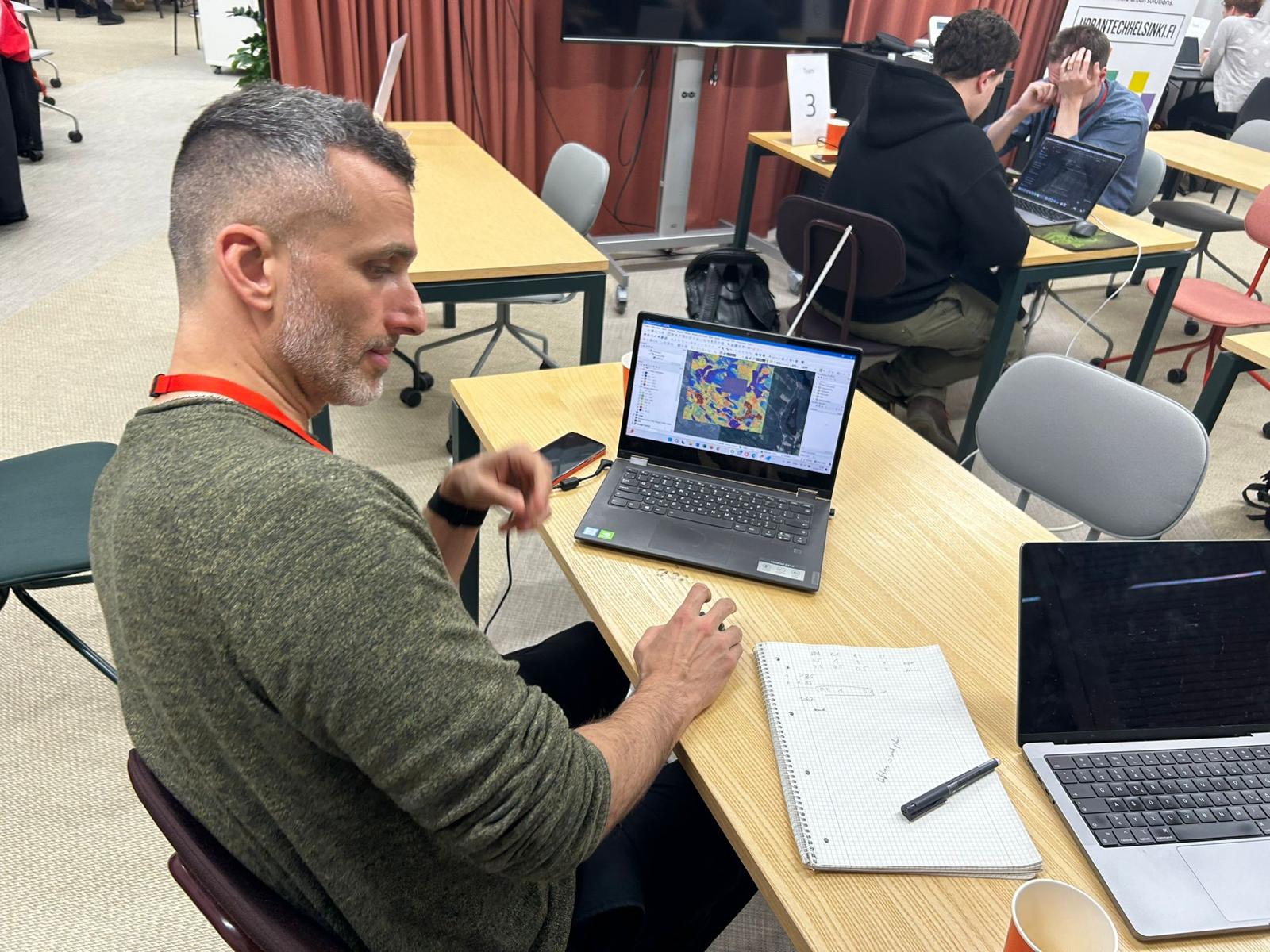 A team member working at a table during the hackathon, with a laptop showing a colourful map and a notebook with handwritten notes beside it.