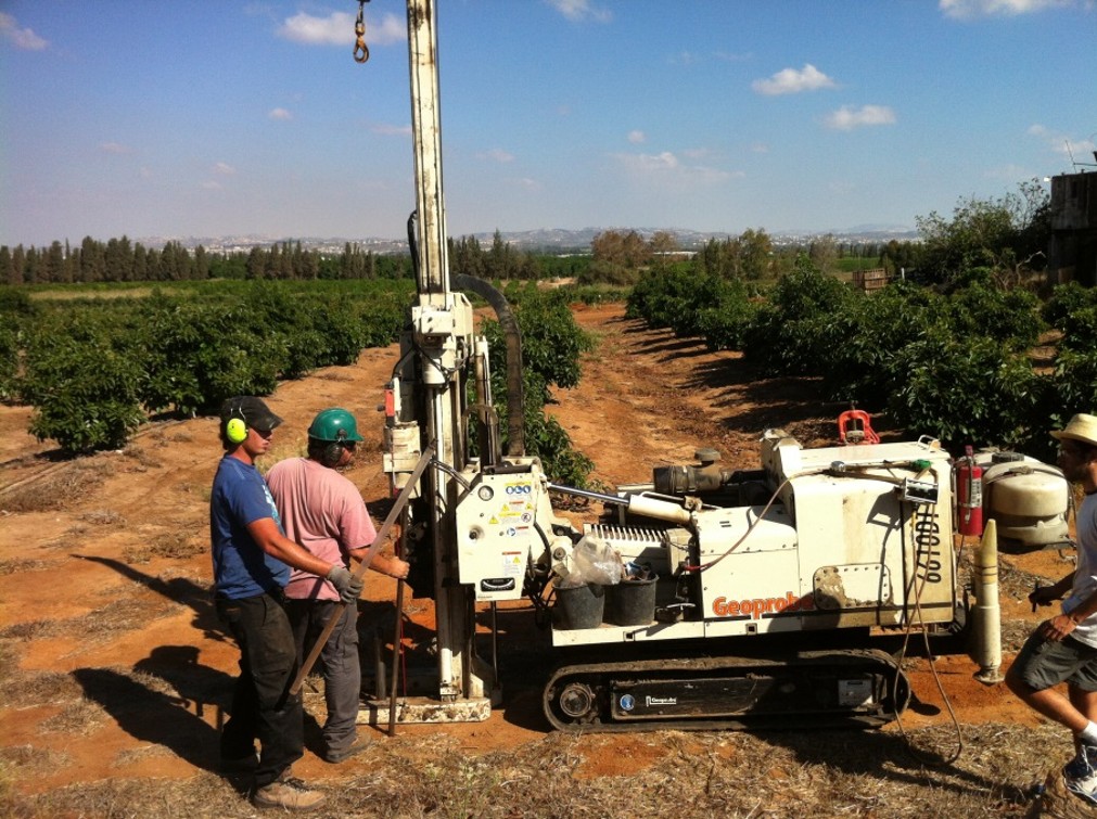 Crew operating a tracked Geoprobe soil-coring rig between rows of trees in an orchard, with the drill mast raised above the ground.